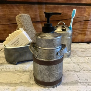 May include: Silver and brown metal soap dispenser shaped like a milk can with a pump top. The dispenser is sitting on a white brick surface.