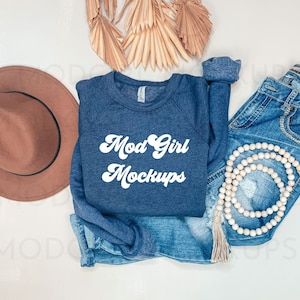 以下が含まれることがあります： 白い文字で「Mod Girl Mockups」と書かれたネイビーブルーのスウェットシャツ。スウェットシャツは、白い背景に、茶色の帽子、青いジーンズ、木製のビーズネックレス、タッセルと一緒に置かれています。