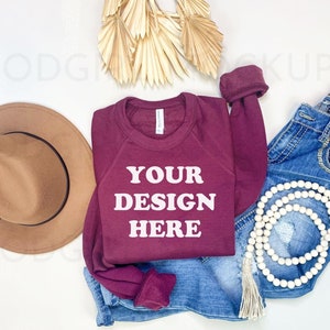 May include: A maroon sweatshirt with the text "YOUR DESIGN HERE" in white. The sweatshirt is laid out on a white surface with a brown hat, blue jeans, a white beaded necklace, and a dried palm leaf.