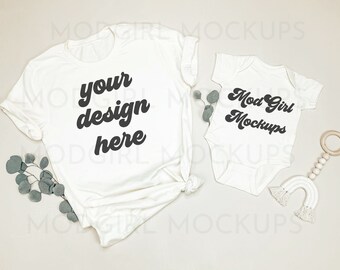 May include: A white t-shirt and a white baby onesie with the text "your design here" and "Mod Girl Mockups" on them. The onesie is lying on a light beige surface with a wooden toy and a white crocheted rainbow.