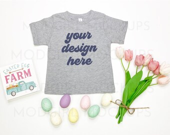 May include: A heather gray t-shirt with the text "your design here" in navy blue. The image includes a sign that says "Easter Egg Farm" and a bouquet of pink and white tulips. Several pastel-colored Easter eggs are also present.