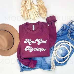 May include: A maroon sweatshirt with the text "Mod Girl Mockups" in white retro font. The sweatshirt is laid out on a white surface with a brown hat, blue jeans, a white beaded necklace, and dried palm leaves.