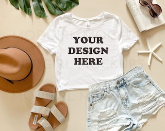 May include: White cropped t-shirt with short sleeves and the text "YOUR DESIGN HERE" printed in black. The t-shirt is laid flat on a beige surface with a brown hat, sandals, denim shorts, a starfish, and a book with sunglasses on top.
