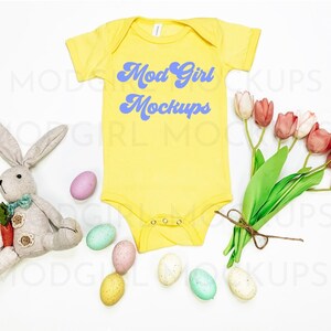 May include: A yellow baby onesie with the text "Mod Girl Mockups" in blue. The onesie is surrounded by pastel Easter eggs and a bunch of pink tulips.