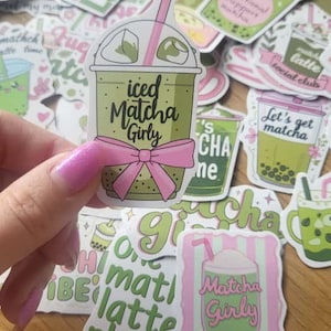 May include: A selection of matcha-themed stickers, with a focus on iced matcha drinks. One sticker is held up, featuring a green iced matcha drink with a pink bow and the text "iced Matcha Girly". Other stickers include phrases like "Matcha Latte" and "Let's get matcha".