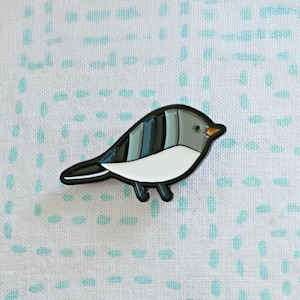 May include: A bird-shaped enamel pin with a black outline. The bird has a white belly, gray head, and a back with blue and gray stripes. The bird's beak is orange. The pin is on a white background with a blue dotted pattern.