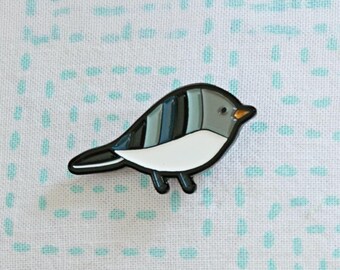 Puede incluir: Un pin de esmalte con forma de pájaro con un contorno negro. El pájaro tiene un vientre blanco, una cabeza gris y una espalda con rayas azules y grises. El pico del pájaro es naranja. El pin está sobre un fondo blanco con un patrón de puntos azules.