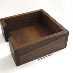 K&ouml;nnte beinhalten: Ein dunkelbrauner, quadratischer Serviettenhalter aus Holz mit einer offenen Vorderseite. Der Halter ist aus massivem Holz gefertigt und hat eine glatte Oberfl&auml;che. Der Text "CubeWoodenDesign" ist in der unteren rechten Ecke des Halters sichtbar.
