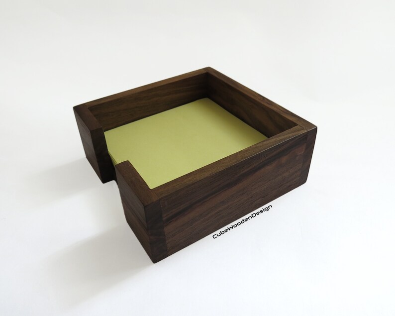 K&ouml;nnte beinhalten: Ein dunkelbrauner Notizblockhalter aus Holz mit einem Schlitz zum Halten von Haftnotizen. Der Halter ist aus Walnussholz gefertigt und hat eine nat&uuml;rliche Oberfl&auml;che. Der Text "CubeWoodenDesign" ist in der unteren rechten Ecke des Halters sichtbar.