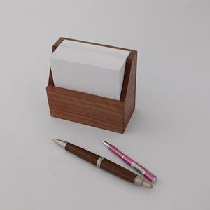 Puede incluir: Un organizador de escritorio de madera con una pila de papel de notas blanco en su interior. Dos bolígrafos, uno marrón y plateado, y un bolígrafo rosa, están sobre la superficie junto al organizador. El organizador y los bolígrafos están sobre una superficie blanca.