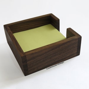 K&ouml;nnte beinhalten: Ein dunkelbrauner Holz-Notizblockhalter mit einem hellgr&uuml;nen Haftnotizblock im Inneren. Der Halter hat eine kleine &Ouml;ffnung an der Seite f&uuml;r einfachen Zugriff auf die Notizen. Der Text "CubeWoodenDesign" ist auf der Unterseite des Halters sichtbar.