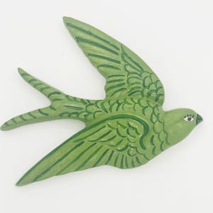 May include: A wall-mounted swallow bird ornament crafted in a light green hue with darker green painted feather details. The bird is depicted in a flying position with wings extended and a forked tail, featuring a small eye with painted eyelashes.