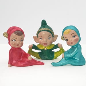May include: Three ceramic elf figurines in red, green, and blue outfits. Each elf has a painted face and a glittery accent on their hat. The elves are in seated poses with their legs spread.