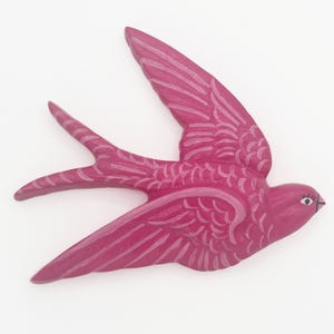 May include: A bright pink swallow wall ornament featuring carved feather details and a painted eye. The bird is depicted in a flying position with wings extended and a forked tail. The surface has a glossy finish with lighter pink accents.