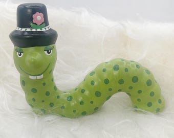 May include: A quirky, green ceramic worm figure with dark green spots. The worm is wearing a black top hat decorated with a pink flower. The worm has a smiling face with white teeth and is posed on a white surface.