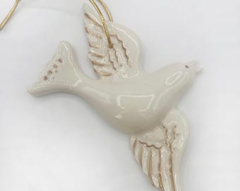 May include: A white ceramic dove ornament with gold accents on the wings. The dove is in flight with its wings spread out. The ornament has a gold string attached to the top for hanging.