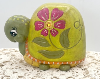 May include: A whimsical, hand-painted ceramic turtle figurine. The turtle features a light green head and legs, with a yellow-green shell adorned with a pink flower design. The shell has a decorative orange border. The turtle is approximately 13 cm long.
