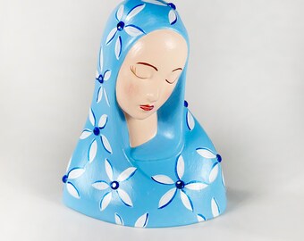 May include: A blue ceramic figurine of a woman with a white floral pattern on her head covering. The figurine has blue eyes and a closed mouth.