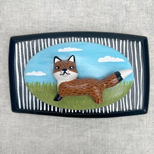 May include: A rectangular, black-framed decorative piece with a painted fox scene. The fox is brown with a white chest and tail tip, set against a blue sky with white clouds and green grass. The frame has white vertical stripes.