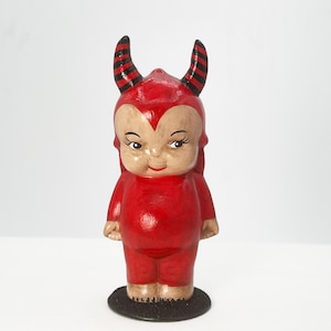 May include: A small figurine of a character wearing a red suit with a hood featuring two black and red striped horns. The character has a pale face, dark eyelashes, and a small red mouth, standing on a circular black base against a white background.