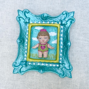 May include: A teal-coloured, ornate picture frame with a small doll inside. The doll wears a green swimsuit with a pink heart and a pink bow. The background has a sky and grass design.