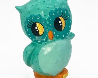 May include: A teal blue owl figurine with white polka dots and large eyes. The owl has a yellow beak and orange feet.