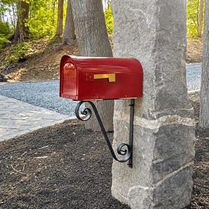 May include: A glossy red metal mailbox with a gold-colored flag attached to the side. The mailbox is mounted onto a tall, rough-hewn gray stone pillar using a black wrought iron bracket featuring decorative scrollwork at the top and bottom.