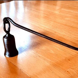 May include: A black metal candle snuffer featuring a long, slender handle with a curved hook at one end and a bell-shaped extinguisher attached to a circular loop at the other end. The object has a smooth, dark finish and a simple, functional design.
