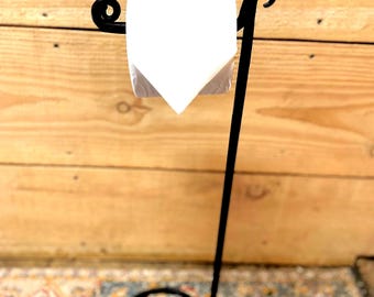 May include: A black metal toilet paper holder featuring a tall, slender vertical stand with a circular base and a horizontal arm with curled ends. A single roll of white toilet paper is mounted on the horizontal arm of the metal stand.