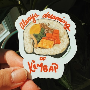 Puede incluir: Una pegatina blanca con una colorida ilustración de kimbap, un plato coreano. La pegatina tiene las palabras "Always dreaming of Kimbap" en escritura roja. La pegatina se sostiene frente a un fondo de hojas verdes.