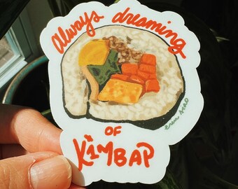 Può includere: Un adesivo bianco con un'illustrazione colorata di kimbap, un piatto coreano. L'adesivo riporta la scritta "Always dreaming of Kimbap" in rosso. L'adesivo è tenuto davanti a uno sfondo di foglie verdi.