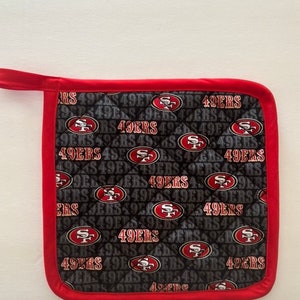 May include: A square, quilted pot holder featuring a repeating pattern of the San Francisco 49ers logo and text on a black background. The pot holder has a red border and a red loop for hanging.