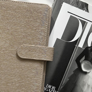 Puede incluir: Una agenda de color topo con una superficie texturizada ondulada y un cierre de correa horizontal. La agenda est&aacute; colocada junto a una revista en blanco y negro con el texto DIOR MAGAZINE y el n&uacute;mero 23 en la portada, mostrando a una persona con vestido.