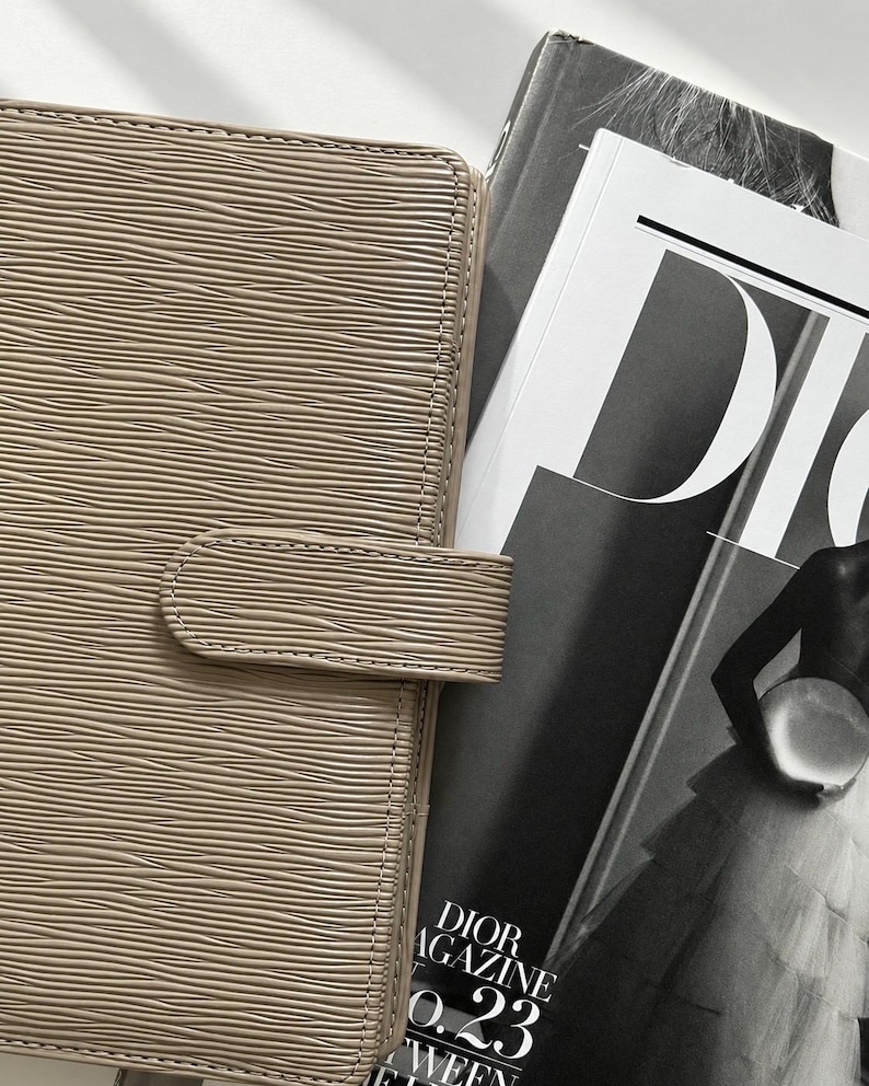 Puede incluir: Una agenda de color topo con una superficie texturizada ondulada y un cierre de correa horizontal. La agenda est&aacute; colocada junto a una revista en blanco y negro con el texto DIOR MAGAZINE y el n&uacute;mero 23 en la portada, mostrando a una persona con vestido.