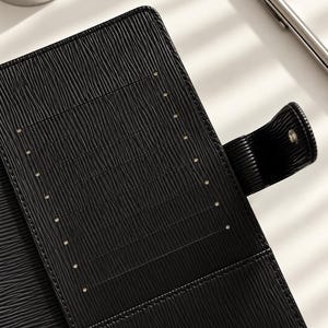 Puede incluir: Una funda de agenda de cuero negro texturizado que presenta una fila vertical de ocho ranuras para tarjetas en el panel interior izquierdo y una correa lateral con cierre de broche met&aacute;lico. El material tiene un patr&oacute;n acanalado horizontal.