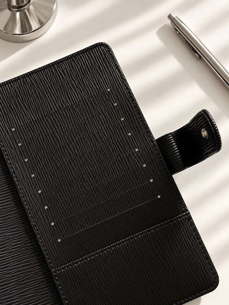 Puede incluir: Una funda de agenda de cuero negro texturizado que presenta una fila vertical de ocho ranuras para tarjetas en el panel interior izquierdo y una correa lateral con cierre de broche met&aacute;lico. El material tiene un patr&oacute;n acanalado horizontal.