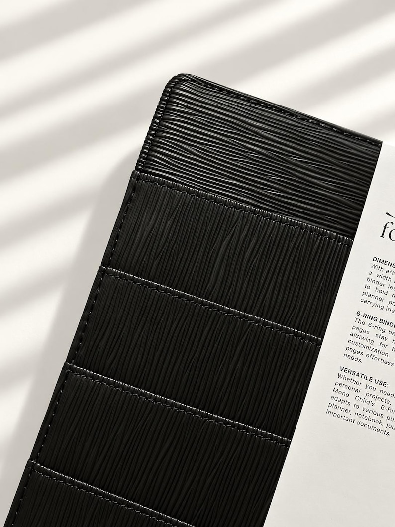 Puede incluir: Una carpeta de 6 anillas negra con un patr&oacute;n de l&iacute;neas horizontales texturizadas en la cubierta. Un inserto de papel blanco est&aacute; unido al lateral, con texto impreso que incluye las palabras DIMENSIONS, 6-RING BINDER y VERSATILE USE, describiendo el producto.
