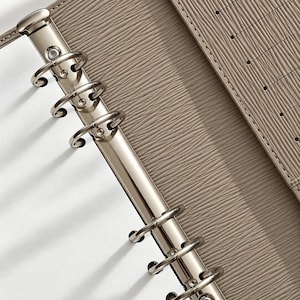 Puede incluir: Una carpeta organizadora de color topo con un mecanismo de seis anillas de metal plateado. La superficie exterior tiene una textura fina con nervaduras horizontales. El panel interior incluye una serie de ranuras verticales para tarjetas.