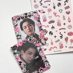 May include: Two photo card holders with a picture of a person, decorated with pink and black stickers. A sheet of stickers with various pink and black designs, including hearts, flames, and text, is also visible. The stickers have the text "MAKNAE", "ANNYEONG!", and "MAKNAE".