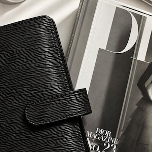 Puede incluir: Una agenda de cuero negro texturizado con cierre de correa horizontal junto a un ejemplar de Dior Magazine No. 23. La agenda presenta un distintivo patr&oacute;n lineal ondulado en relieve en su superficie, acompa&ntilde;ada por un bol&iacute;grafo plateado cerca.