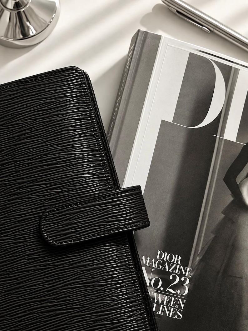 Puede incluir: Una agenda de cuero negro texturizado con cierre de correa horizontal junto a un ejemplar de Dior Magazine No. 23. La agenda presenta un distintivo patr&oacute;n lineal ondulado en relieve en su superficie, acompa&ntilde;ada por un bol&iacute;grafo plateado cerca.