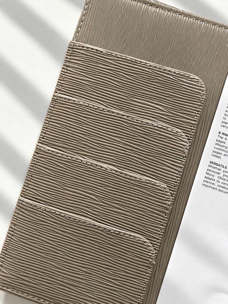 Puede incluir: Una funda para agenda de color topo con un patr&oacute;n de l&iacute;neas onduladas texturizadas y tres ranuras horizontales para tarjetas en la parte frontal. La funda est&aacute; parcialmente abierta, revelando una p&aacute;gina blanca con texto impreso que incluye 6-RING y VERSATILE.