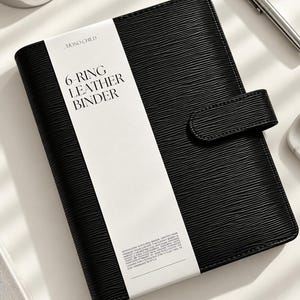 Puede incluir: Una carpeta de cuero negro de 6 anillas con una superficie texturizada y una correa de cierre magn&eacute;tico. Una banda de papel blanca envuelve el centro, con el texto MONO CHILD y 6-RING LEATHER BINDER en negro, junto con texto impreso peque&ntilde;o.