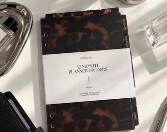 Puede incluir: Un juego de separadores de agenda de 12 meses con un patrón de carey marrón y negro. Los separadores tienen pestañas negras etiquetadas con los trimestres Q1 a Q4 y los meses de enero a junio. Una banda de papel blanca muestra el texto MONO CHILD 12-MONTH PLANNER DIVIDERS A5 SIZE DESIGNED TO HELP YOU PLAN WITH INTENTION.
