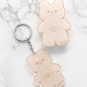 Op de afbeelding: Twee beige beren-sleutelhangers en een bijpassende bedel. Het berenontwerp heeft een hartvormige neus en de tekst "you're gonna be happy" en "00:00". Eén sleutelhanger heeft een zilveren sleutelring en ketting.