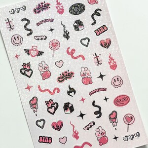 May include: Sheet of stickers with a glittery background. The stickers feature pink and black designs, including hearts, flames, a bunny, and text that reads "BIAS", "ANNYEONG!", and "MAKNAE". The stickers are in various shapes and sizes.