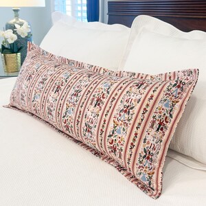 May include: A long, rectangular decorative pillow with a cream background and vertical stripes. The stripes are accented with red and feature a repeating pattern of figures and floral elements. The pillow is placed on a white bed.