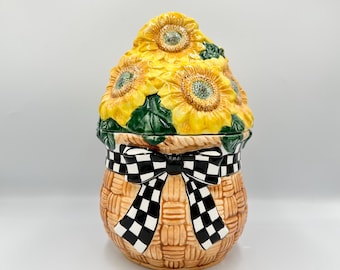 Op de afbeelding: Een keramieken koekjespot in de vorm van een gevlochten mand met een deksel voorzien van gebeeldhouwde gele zonnebloemen en groene bladeren. De mand heeft een beige, getextureerde afwerking en een zwart-wit geblokte strik.