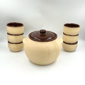 May include: A ceramic serving set consisting of a large lidded pot and six small bowls. The pieces feature a two-tone design with a matte cream-colored exterior and a glossy dark brown glaze on the pot lid and the interior of all vessels.