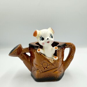 May include: A ceramic watering can planter in a rich brown color, featuring a charming white puppy figurine peeking out. The watering can has embossed floral and sunburst designs, adding a vintage touch to this decorative piece.